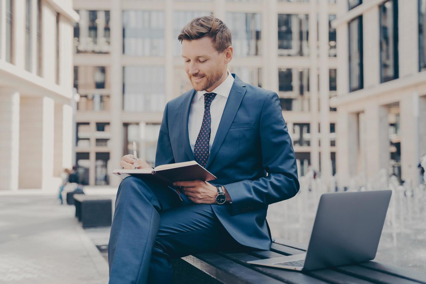 relaxed-confident-office-worker-in-formal-blue-suit-writing-notes-in-agenda-while-working-outside-free-photo